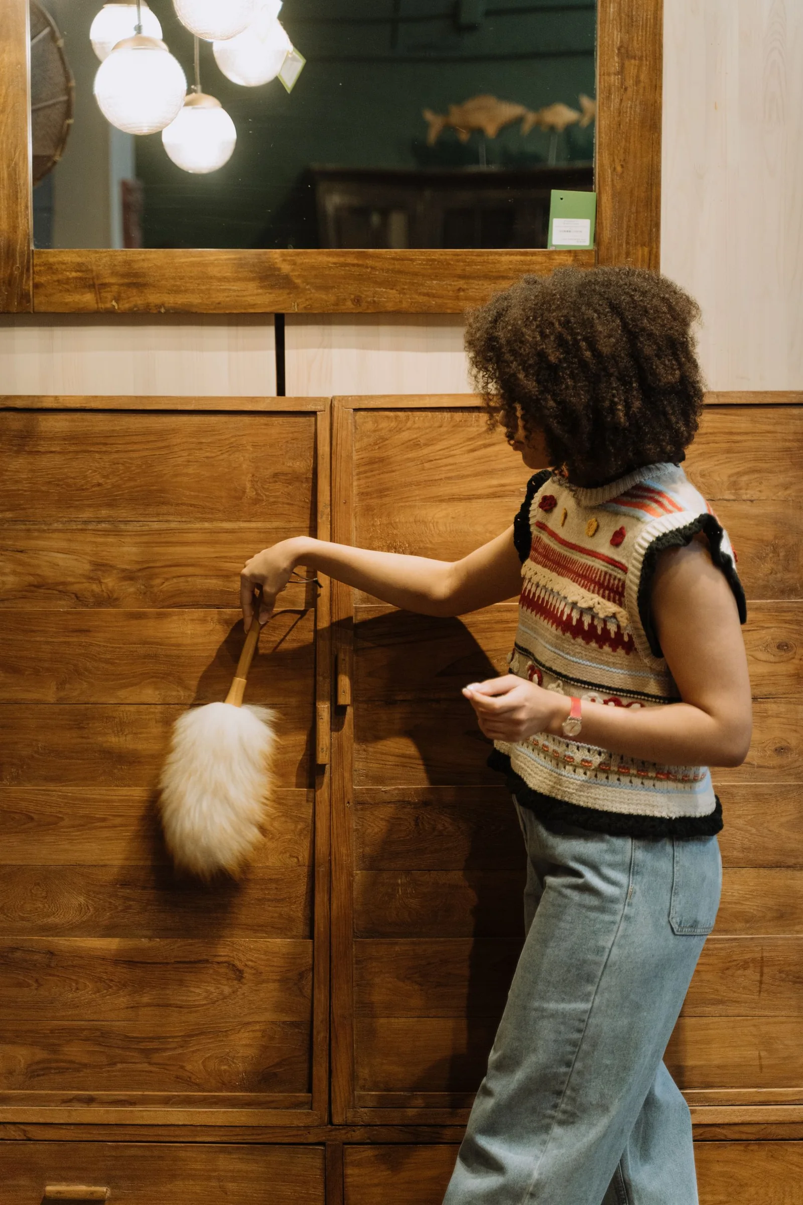 A casually dressed woman uses a fluffy duster on a large wooden cabinet beneath a mirror.