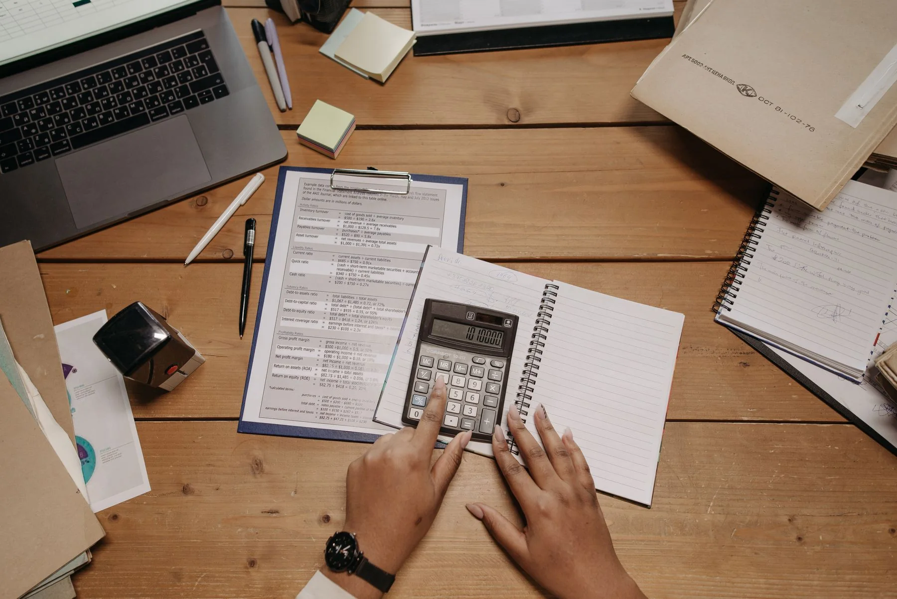 Overhead view of hands using a calculator beside paperwork, a notebook and a laptop on a wooden desk.
