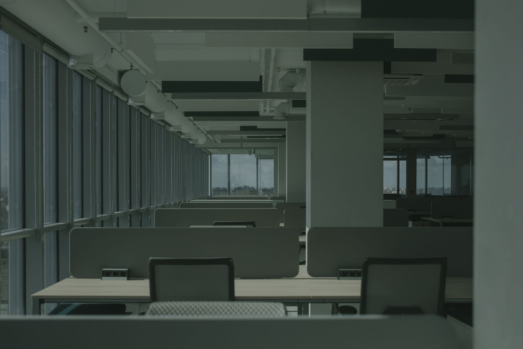 Rows of desks and mesh office chairs beside tall windows in an empty open-plan office.