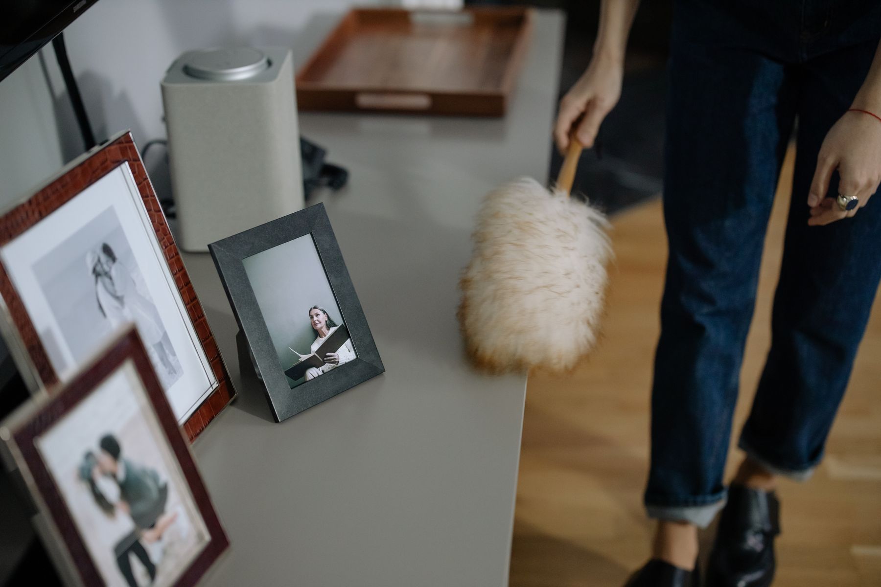 A hand moves a fluffy duster along a gray shelf holding framed photographs.