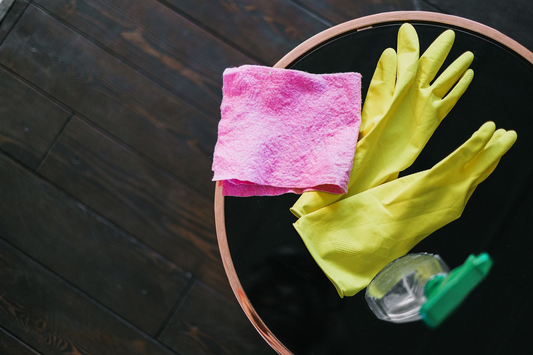 Yellow rubber gloves, a pink cleaning cloth and a green-trigger spray bottle on a round black table.