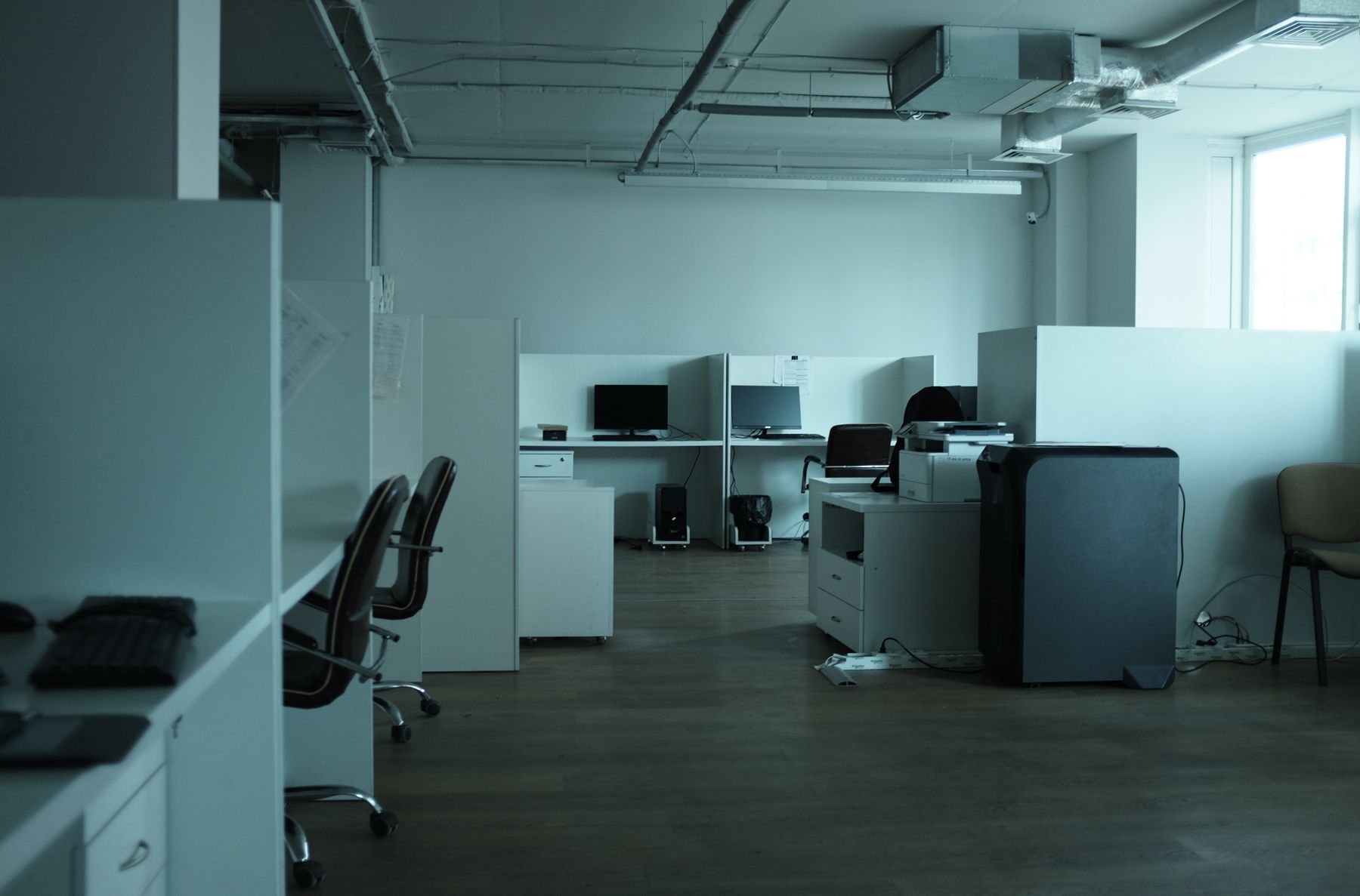 Unoccupied office with desks, computer monitors, swivel chairs and a printer beneath exposed ceiling pipes.