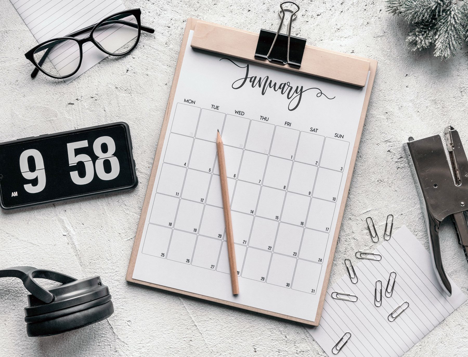 January calendar on a clipboard beside a digital clock, glasses, headphones and office stationery.