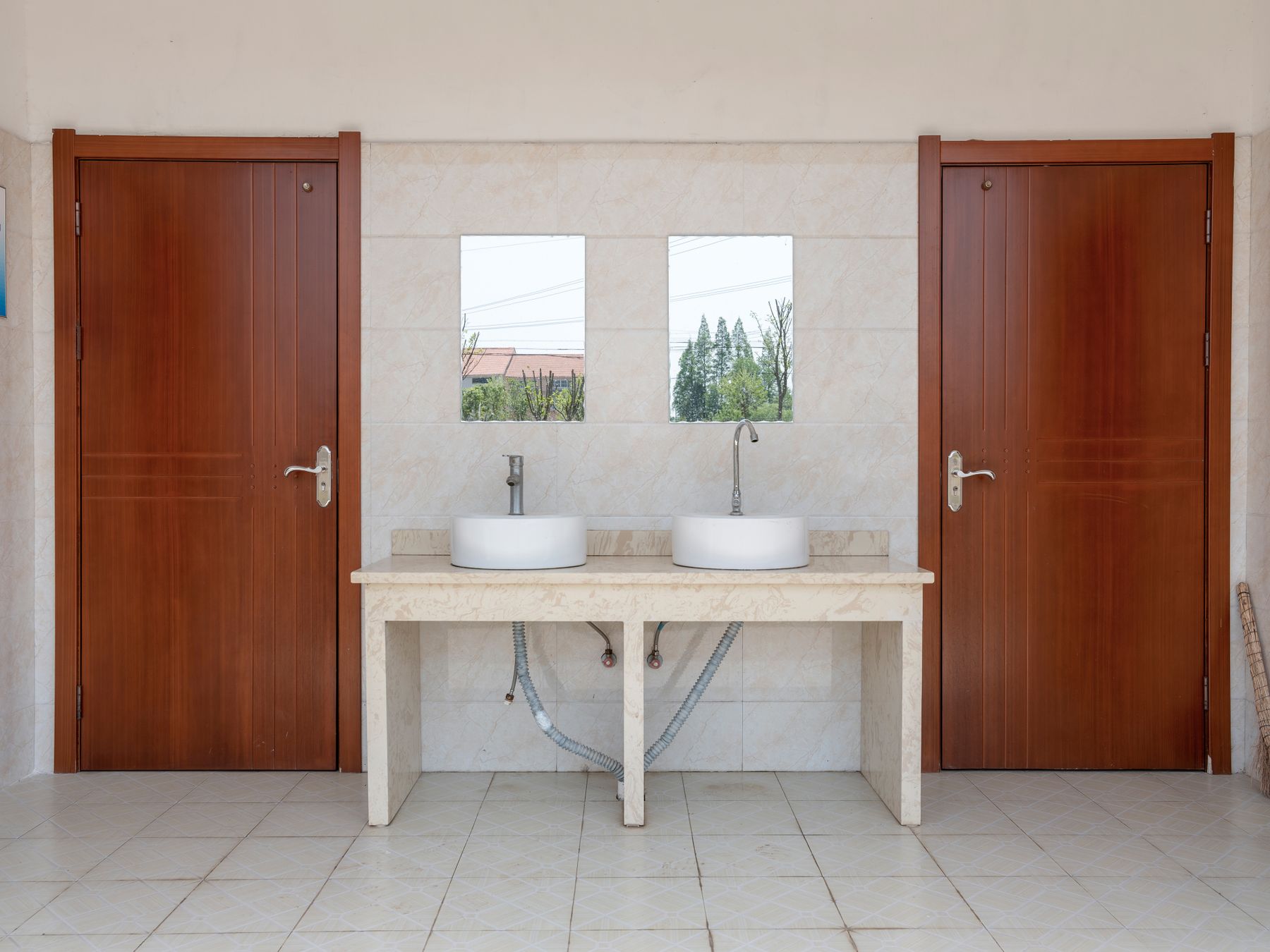 Two white vessel sinks and mirrors between wooden restroom doors above a tiled floor.