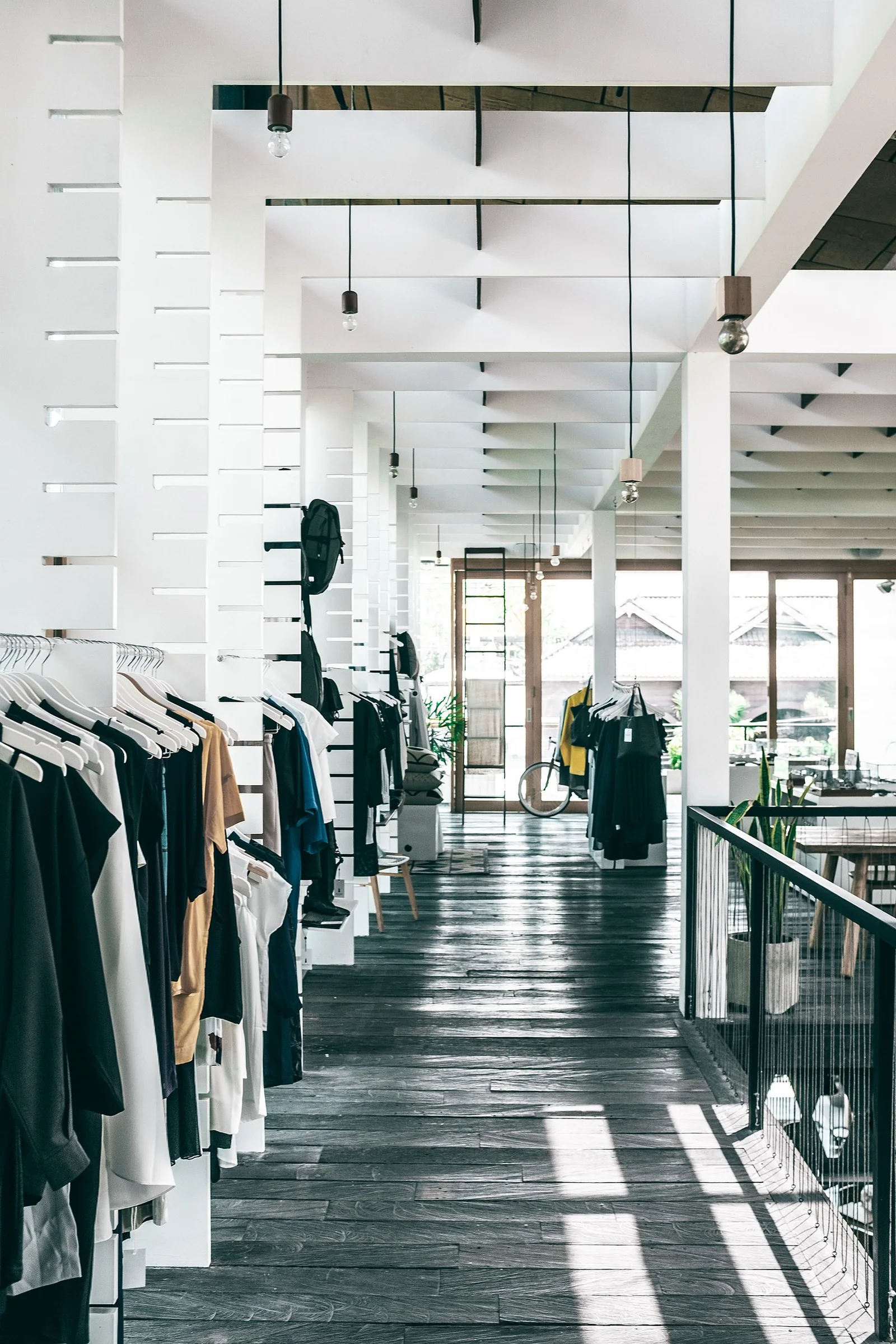 Sunlit clothing store aisle with garment racks, mirrors and a reflective dark floor.